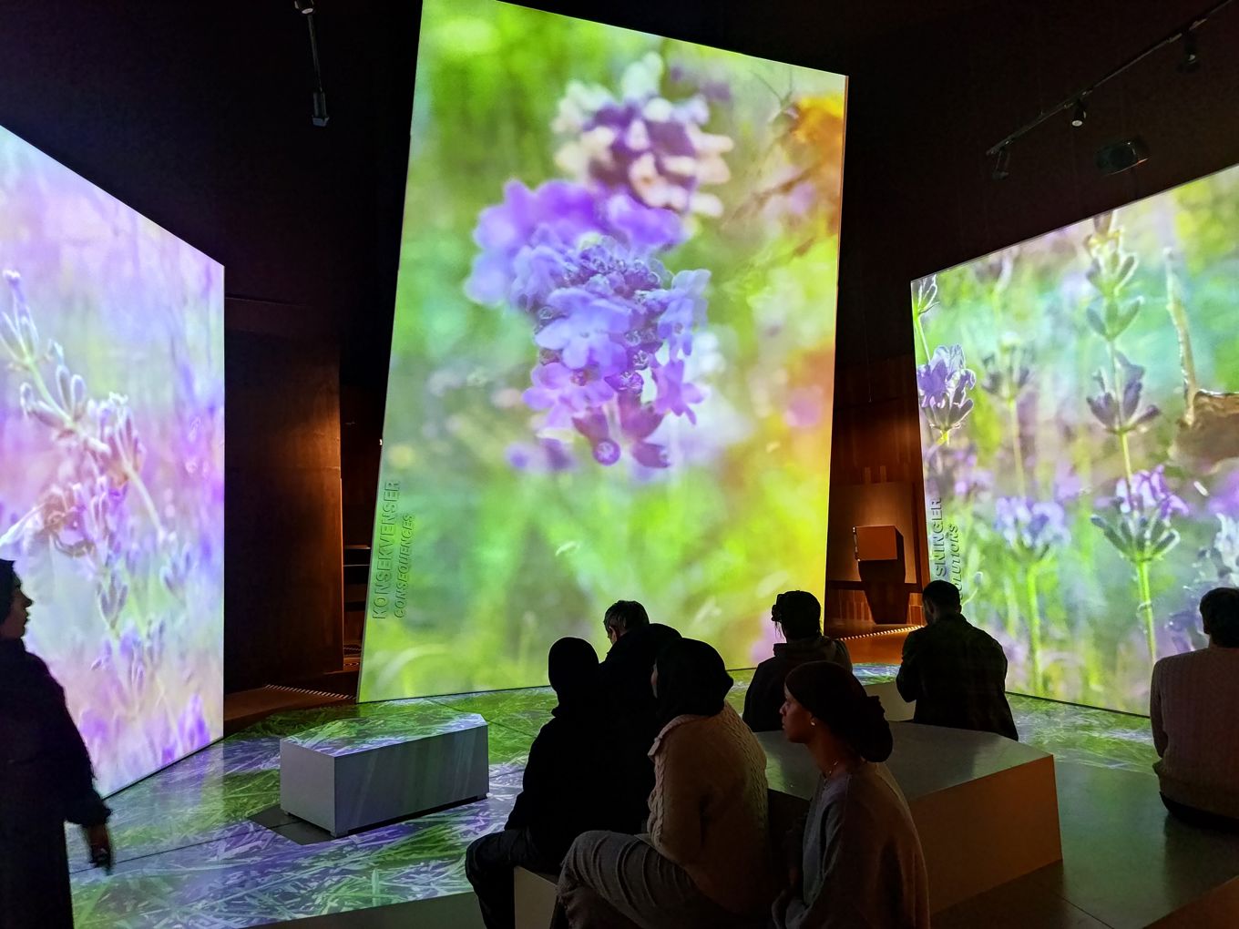 en gruppe mennesker som sitter i et rom med en scene og en stor skjerm med blomster på